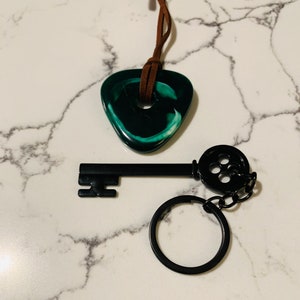 May include: A black key with a round, black button detail on the end of the key. The key is attached to a black key ring. A green and white swirl pendant is attached to a brown leather cord.