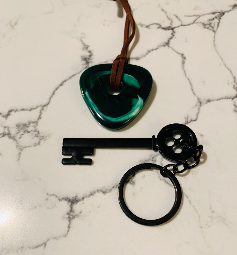 May include: A black key with a round, black button detail on the end of the key. The key is attached to a black key ring. A green and white swirl pendant is attached to a brown leather cord.