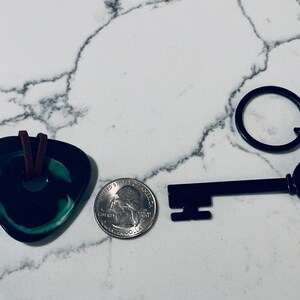 May include: A black metal key with a round keyhole and a small chain attached to a key ring. The key is next to a green glass pendant with a brown leather cord.