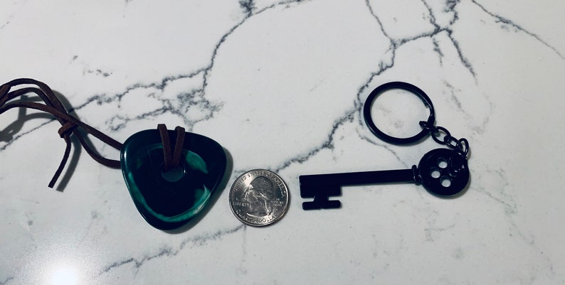 May include: A black metal key with a round keyhole and a small chain attached to a key ring. The key is next to a green glass pendant with a brown leather cord.