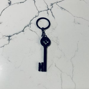 May include: A black metal key with a round button-like head and a key ring attached. The key is on a white marble background.