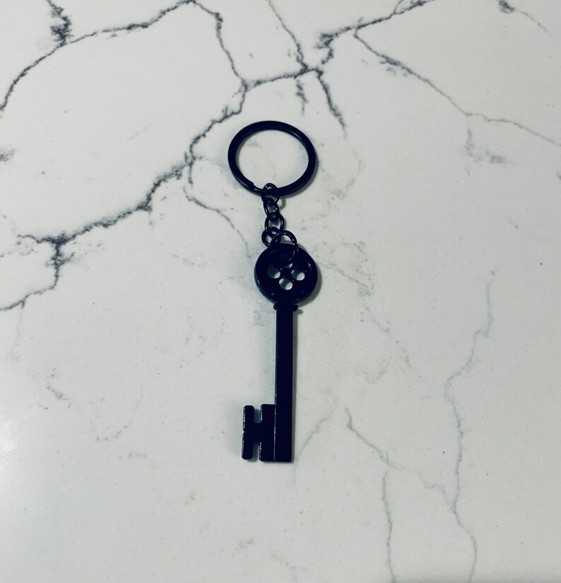 May include: A black metal key with a round button-like head and a key ring attached. The key is on a white marble background.