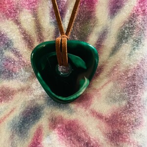May include: A green and white stone pendant with a hole in the center, hanging from a brown leather cord. The pendant is shaped like a teardrop and has a swirl pattern.
