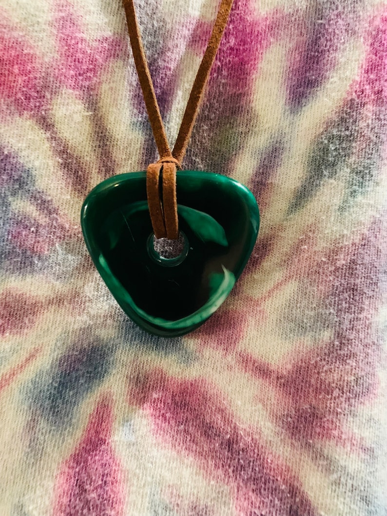 May include: A green and white stone pendant with a hole in the center, hanging from a brown leather cord. The pendant is shaped like a teardrop and has a swirl pattern.