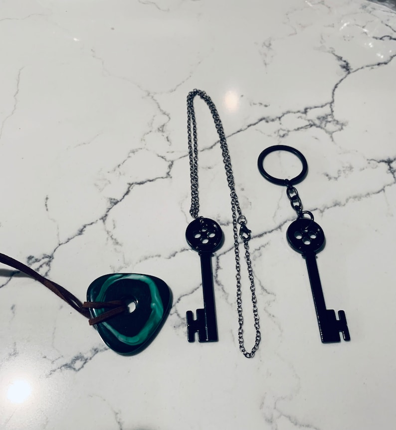 May include: Three necklaces with key pendants. One necklace has a black key pendant with a silver chain. Another necklace has a black key pendant with a silver chain and a silver key ring. The third necklace has a green and black stone pendant with a brown leather cord.