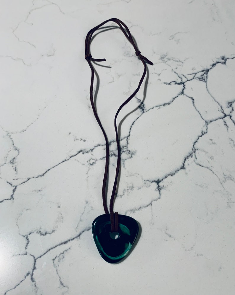 May include: A dark green, triangular pendant with a hole in the center hangs from a brown leather cord. The pendant has a swirl pattern.