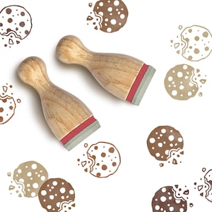 Könnte beinhalten: Zwei holzfarbene Keksstempel mit roten und grauen Basen werden auf weißem Hintergrund gezeigt, umgeben von Keksillustrationen in verschiedenen Brauntönen. Die Stempel haben eine natürliche Holzoberfläche und sind für Bastelarbeiten oder zum Backen gedacht.