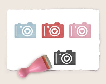 May include: Four rubber stamps with a camera design and a heart in the lens. The stamps are in blue, red, pink, and black. A pink wooden handle is included.