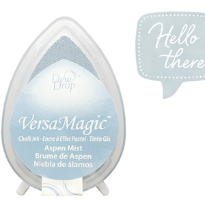 May include: A light blue ink pad with the text "Dew Drop VersaMagic Chalk Ink - Encre à Effet Pastel - Tinta Gis Aspen Mist Brume de Aspen Niebla de álamos" on the front. The ink pad is in the shape of a teardrop and is sitting on a white stand. A speech bubble with the text "Hello there" is in the upper right corner of the image.