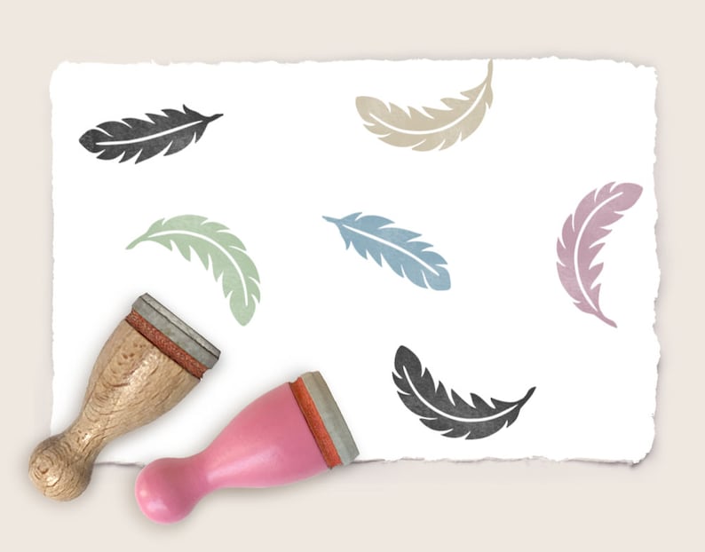 K&ouml;nnte beinhalten: Ein Satz von sechs Holzstempeln mit verschiedenen farbigen Federdesigns. Die Stempel befinden sich auf einem wei&szlig;en Papierhintergrund. Zwei der Stempel sind mit ihren Holzgriffen abgebildet.