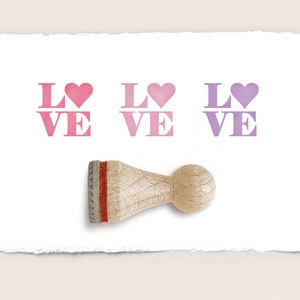 Può includere: Tre timbri in gomma con la parola "LOVE" in rosa e viola con un cuore sopra la "O". I timbri sono su un foglio di carta bianco con un manico in legno.