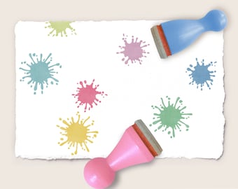 May include: A white paper with colorful paint splatters in yellow, pink, green, and blue. Two rubber stamps with wooden handles, one blue and one pink, are positioned near the paper, suggesting a craft or art project.