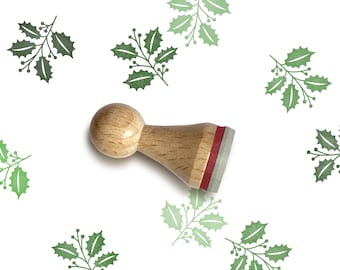 May include: A wooden stamp with a red and gray ink pad. The stamp is shaped like a small, round, wooden handle with a rectangular base. The stamp is used to create a green holly leaf design.