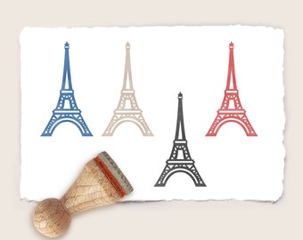 May include: Four rubber stamps featuring a silhouette of the Eiffel Tower in different colors: blue, beige, red, and black. The stamps are on a white background with a torn edge.