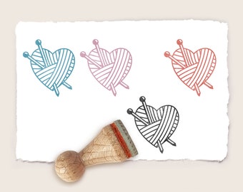 May include: A wooden stamp with a heart-shaped design featuring yarn and knitting needles. The image shows four heart designs in blue, pink, red, and black. The stamp has a wooden handle.