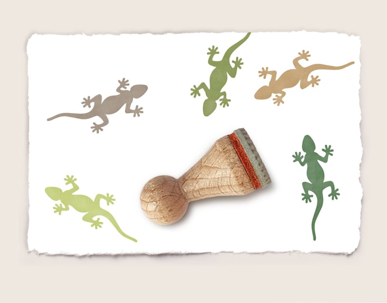 K&ouml;nnte beinhalten: Ein Stempel aus Holz mit einem braunen Griff und einer roten Gummifl&auml;che. Der Stempel zeigt eine Silhouette eines Eidechsen. Der Stempel ist umgeben von vier gedruckten Bildern von Eidechsen in verschiedenen Farben, darunter Gr&uuml;n, Braun und Beige.