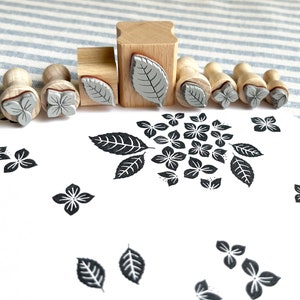 May include: A set of nine wooden stamps with different floral and leaf designs. The stamps are arranged in a row on a striped fabric background. The stamps are made of wood and have a rubber surface. The designs are in black and white.