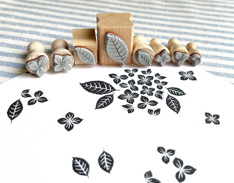 May include: A set of nine wooden stamps with different floral and leaf designs. The stamps are arranged in a row on a striped fabric background. The stamps are made of wood and have a rubber surface. The designs are in black and white.