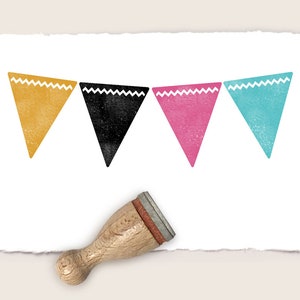 Puede incluir: Un sello de banner colorido con un mango de madera. El banner presenta cuatro banderas triangulares en oro, negro, rosa y turquesa, cada una con un borde en zigzag blanco. El sello está sobre papel blanco texturizado.