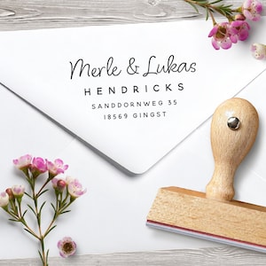 Puede incluir: Un sobre blanco con los nombres "Merle & Lukas" impresos en cursiva, junto con una dirección. Un sello de madera con una almohadilla de tinta roja y blanca está al lado del sobre. Flores rosas están esparcidas alrededor del sobre.