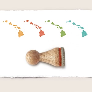 Può includere: Un timbro in legno con un inserto in gomma rossa, utilizzato per creare un'impronta dello stato delle Hawaii. Il timbro è mostrato su uno sfondo di carta bianca con quattro impronte dello stato delle Hawaii di diversi colori.