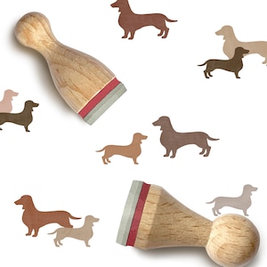 Puede incluir: Dos sellos de madera con diseño de perro salchicha, con detalles en rojo y gris, junto a siluetas de perros salchicha en tonos marrones y beige. Los sellos tienen un mango redondeado y una base rectangular.