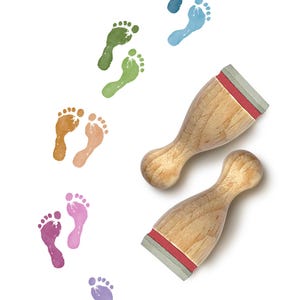 May include: Two wooden stamps with light-coloured grain and red foam bases, positioned next to a series of colourful, stamped footprint patterns in shades of blue, green, orange, pink, and purple, arranged in a trail on a white background.