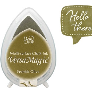 May include: A clear plastic ink pad with a green ink pad inside. The ink pad is labeled "Dew Drop VersaMagic Multi-surface Chalk Ink Spanish Olive". The ink pad is on a white background. A speech bubble with the text "Hello there" is in the upper right corner of the image.