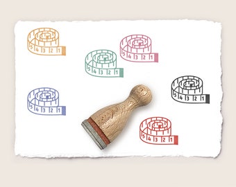 May include: Five rubber stamps with a design of a tape measure with numbers 1 through 15. The stamps are in different colors: orange, green, pink, black, and red. A wooden stamp handle is also shown.