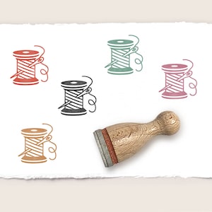 Può includere: Un timbro di legno con un disegno di bobina di filo rosso, arancione, verde e nero. Il timbro è su uno sfondo di carta bianca.
