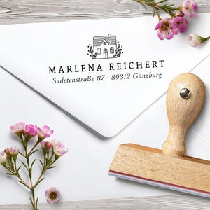 Könnte beinhalten: Ein Holzstempel mit einem Hausdesign, der auf einem weißen Umschlag liegt. Der Umschlag trägt den Namen "MARLENA REICHERT" und Adressdetails. Rosa Blumen und Grünzeug sind um den Umschlag herum angeordnet.