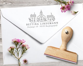 May include: A wooden stamp with a house and church design, resting on a white envelope. The stamp has a wooden handle and a red and white ink pad. The envelope has the name "Bettina Lindemann" and address printed on it. Pink flowers are in the background.