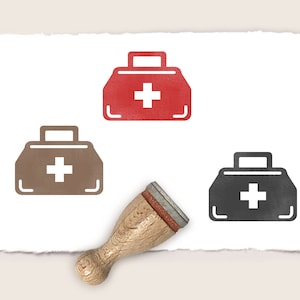 K&ouml;nnte beinhalten: Drei Stempel aus Gummi mit einem Erste-Hilfe-Kasten-Motiv. Die Stempel sind rot, braun und schwarz. Der Erste-Hilfe-Kasten hat ein wei&szlig;es Kreuz. Die Stempel befinden sich auf einem wei&szlig;en Blatt Papier mit einem rauen Rand.