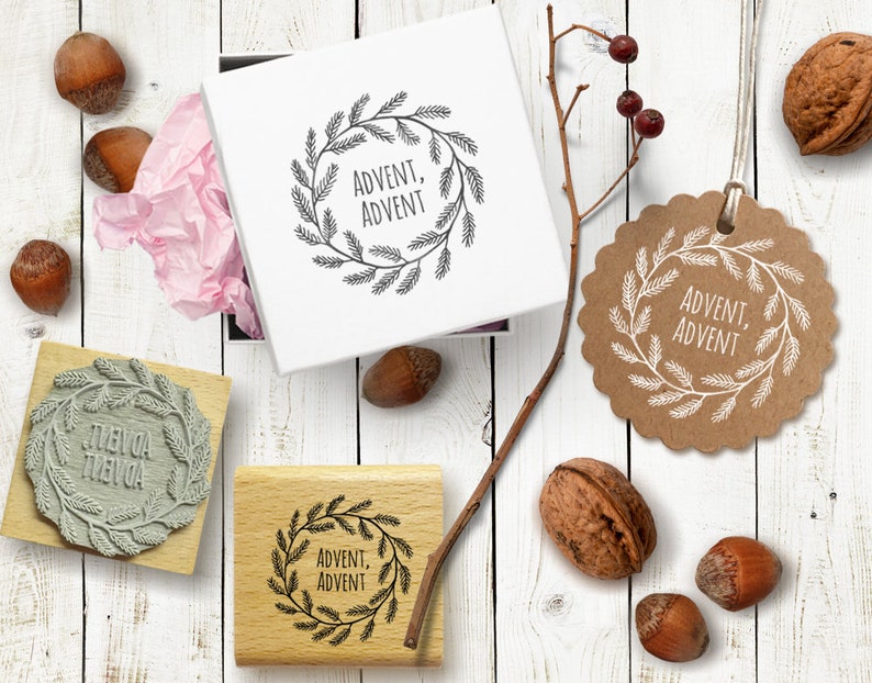 Op de afbeelding: Vier houten stempels met een kransontwerp en de tekst "Advent, Advent" in zwarte inkt. De stempels liggen op een wit houten oppervlak met hazelnoten en walnoten.