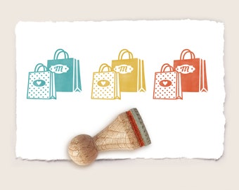 May include: Four different colored rubber stamps of shopping bags with a heart and the letter "M" on the tag. The stamps are on a white piece of paper with a wooden handle.
