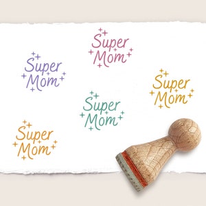 Könnte beinhalten: Ein Stempel aus Holz mit einem Griff und einer rechteckigen Basis. Der Stempel wird verwendet, um ein Design zu erstellen, das "Super Mom" mit Sternen um den Text sagt. Der Stempel befindet sich auf einem weißen Blatt Papier mit vier weiteren Abdrücken desselben Designs in verschiedenen Farben.