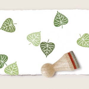 Könnte beinhalten: Ein Stempel aus Holz mit einem roten und grünen Stempelkissen, mit dem man Blattmotive erstellen kann. Der Stempel befindet sich auf einem weißen Blatt Papier mit mehreren grünen Blattabdrücken.