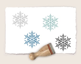 May include: Four snowflake stamps, one in gray, one in blue, one in teal, and one in black, on a white paper background. A wooden stamp handle is in the lower right corner of the image.