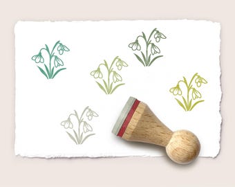 May include: A wooden rubber stamp with a round handle and a red cushion layer, positioned on a piece of textured white paper. The paper features four stamped illustrations of snowdrop flowers in varying shades of green, teal, and light gray.