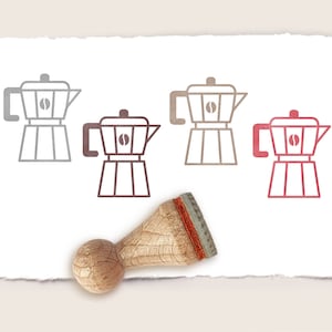Può includere: Tre timbri in gomma con disegni di colore diverso di una caffettiera con un chicco di caffè all'interno. I timbri sono su un foglio di carta bianco con un manico in legno.