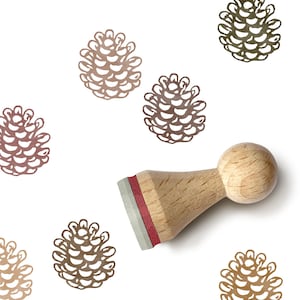 Könnte beinhalten: Ein Holzstempel mit einem Tannenzapfen-Design und einem roten und silbernen Stempelkissen. Mehrere Tannenzapfen-Illustrationen in Braun- und Grüntönen auf weißem Hintergrund.