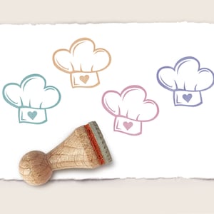 Könnte beinhalten: Vier Stempel aus Gummi mit verschiedenen farbigen Umrissen einer Kochmütze mit einem Herz in der Mitte. Die Stempel befinden sich auf einem weißen Blatt Papier mit einem Holzgriff.