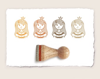 May include: A wooden stamp with a red ink pad features a line of four princess illustrations. Each princess has a crown, long hair, and a simple dress. The illustrations are in a line, with the stamp positioned below the center of the line.