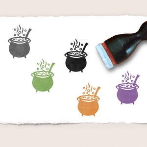 Op de afbeelding: Vijf rubberen stempels met verschillende gekleurde afbeeldingen van een ketel met een houten lepel en stoom die uit de bovenkant komt. De ketel is gevuld met bellen. De stempels bevinden zich op een wit vel papier.