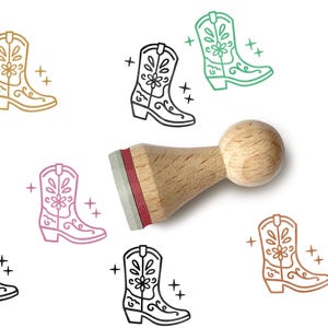 Könnte beinhalten: Ein Stempel mit Holzgriff und rotem Stempelkissen, umgeben von Illustrationen von Cowboy-Stiefeln in Gold, Schwarz, Grün, Rosa und Braun. Jedes Stiefeldesign enthält florale und dekorative Elemente.