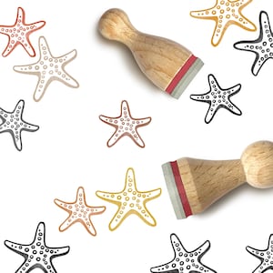 May include: Two wooden stamps with rounded handles and rubber bases are positioned among multiple stamped starfish designs in various colors including black, mustard yellow, and muted orange, scattered across a plain white background.