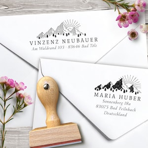 May include: A wooden stamp with a mountain and sun design, next to two white envelopes with printed addresses. The stamp has a light wood handle and a red ink pad. The envelopes have the names and addresses of Vinzenz Neubauer and Maria Huber.
