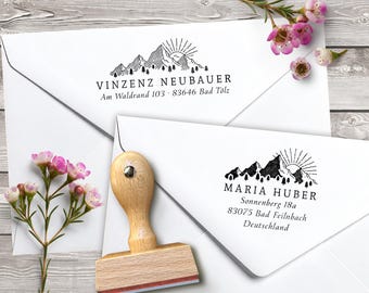 May include: A wooden stamp with a mountain and sun design, next to two white envelopes with printed addresses. The stamp has a light wood handle and a red ink pad. The envelopes have the names and addresses of Vinzenz Neubauer and Maria Huber.