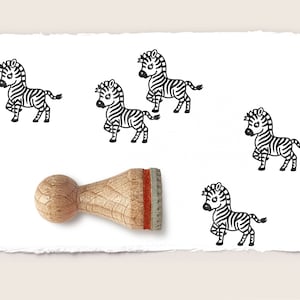 Op de afbeelding: Een houten stempel met een rood en grijs handvat. De stempel heeft een zwart-witte illustratie van een zebra. Er zijn vijf zebra-illustraties op een witte achtergrond.