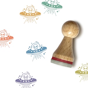 May include: A wooden rubber stamp with a red base and a round wooden handle. The stamp features a line drawing of a flying saucer with an alien inside. The stamp is surrounded by five different colored impressions of the same design.